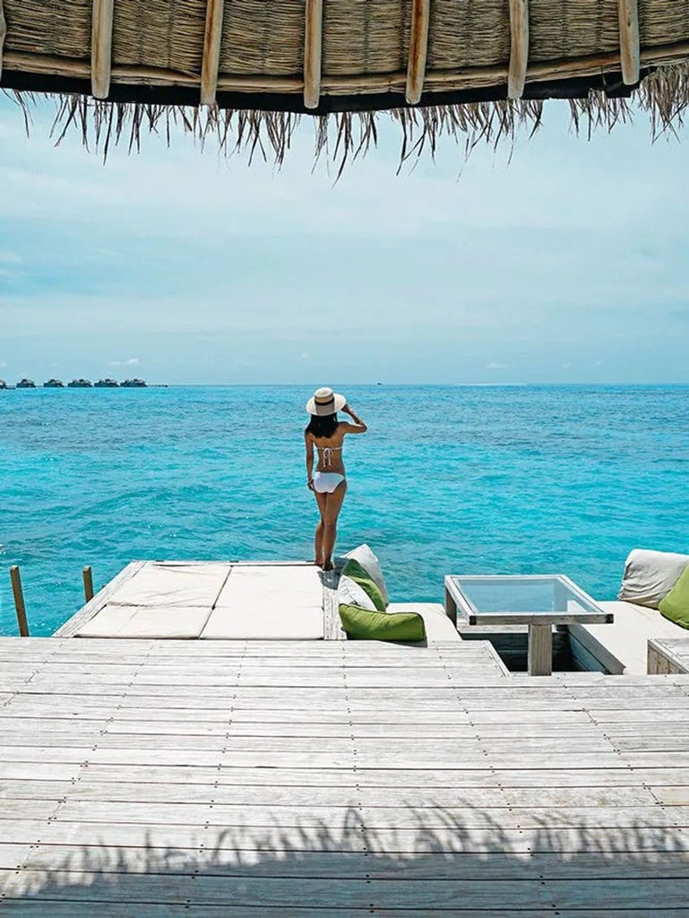 Overwater luxury villa with wooden deck in Maldives, turquoise lagoon below, surrounded by lush tropical vegetation and sandy beach