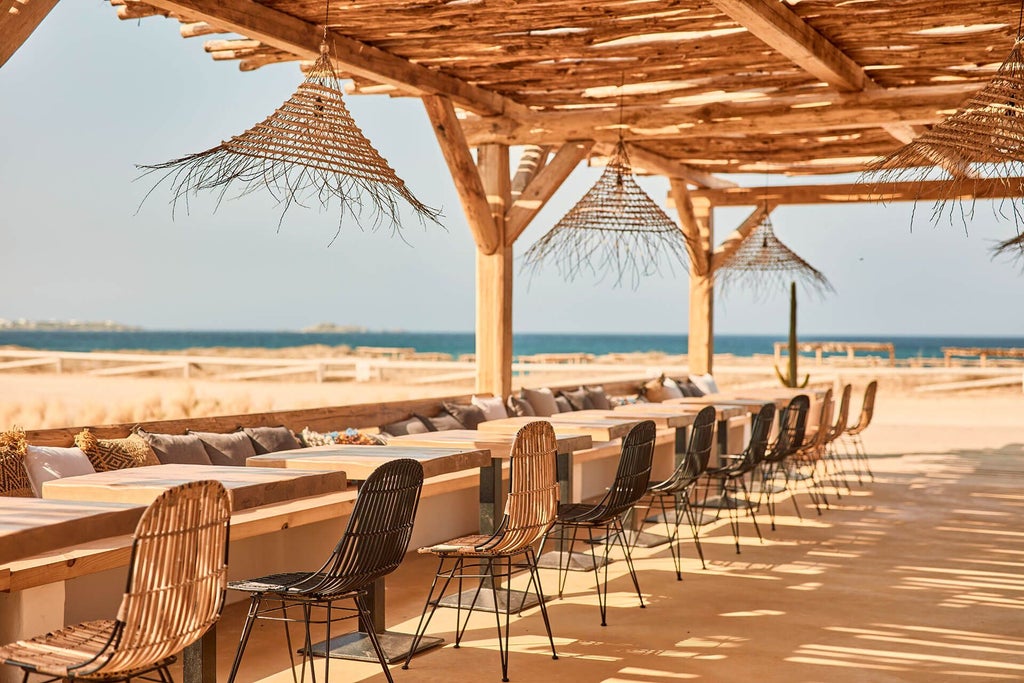 Luxurious beachfront hotel with white minimalist architecture, azure Aegean waters, and sun-drenched terrace overlooking pristine Greek coastal landscape