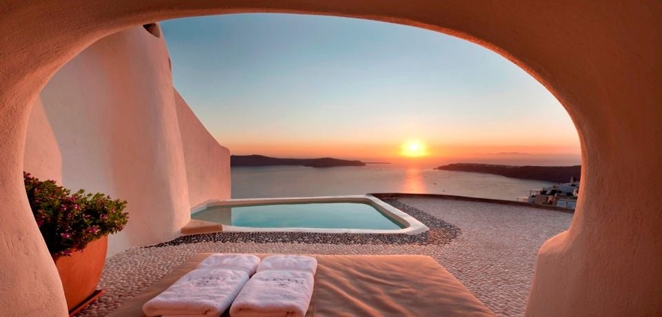 Whitewashed luxury cave suite with private infinity pool overlooking Santorini's caldera and Aegean Sea at sunset, built into cliffside