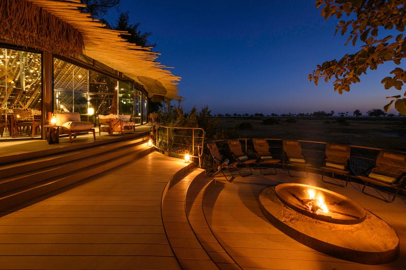 Elevated luxury safari lodge with thatched roof pavilions connected by wooden walkways, overlooking Okavango Delta wetlands at sunset