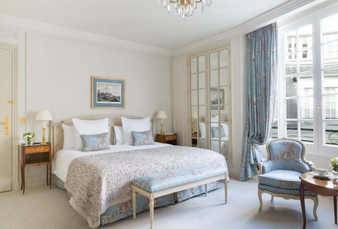 Elegant hotel room with antique French furniture, plush floral curtains, crystal chandelier, king bed, and marble bathroom in soft beige tones