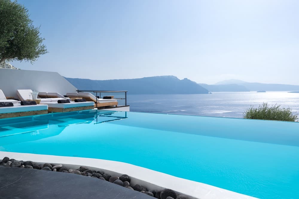 Elegant infinity pool merges with Aegean Sea views, white minimalist loungers line poolside at luxury Santorini resort at sunset