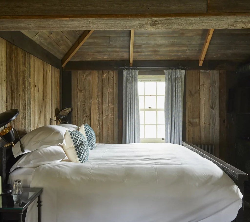 Luxurious forest cabin bedroom at Lime Wood, featuring rustic wood paneling, plush bedding, and large windows overlooking lush woodland scenery.