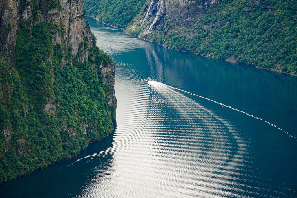 Scenic Norwegian fjord with steep, lush green cliffs, azure waters, and a luxurious cruise boat navigating the dramatic Geirangerfjord landscape