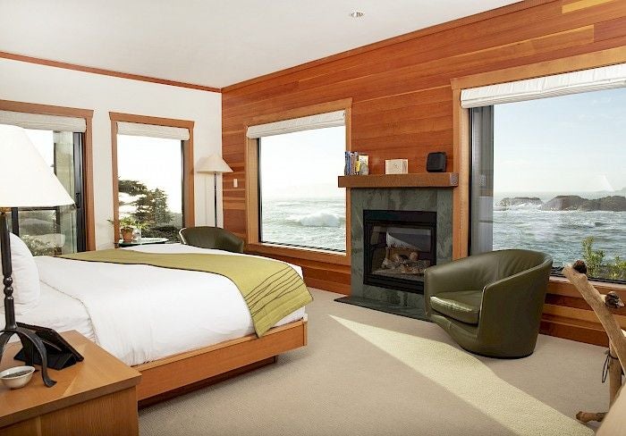 Luxury waterfront lodge with cedar-clad architecture perched on rocky Pacific coast, waves crashing below panoramic windows at sunset