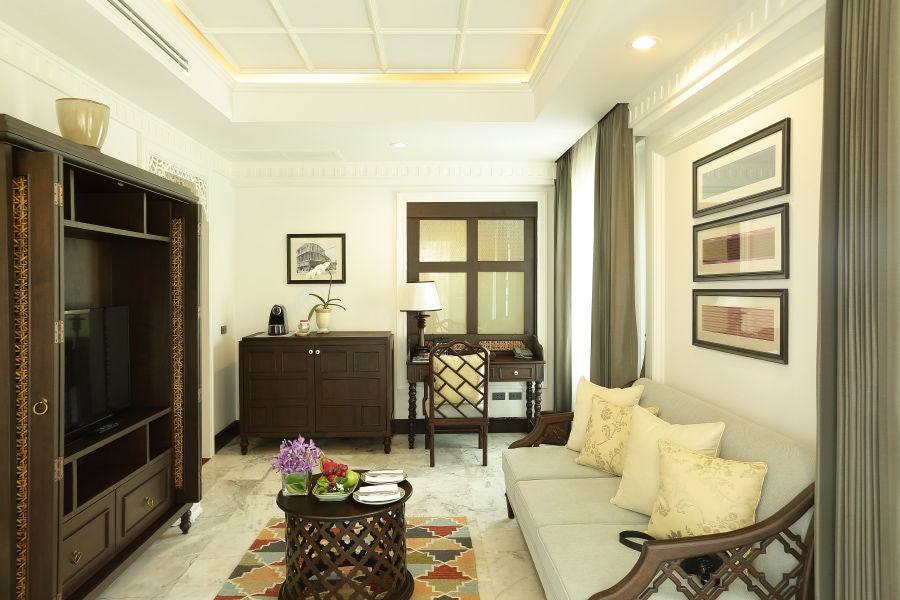 Elegant colonial suite with four-poster bed, ornate wooden furniture, white linen, and traditional Thai-inspired decor in natural light