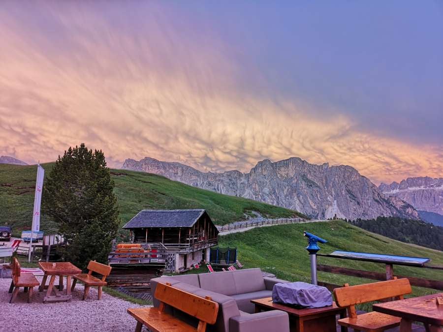 Modern mountain lodge with panoramic glass windows reflecting snow-capped Dolomite peaks, nestled on a scenic Alpine meadow at sunset