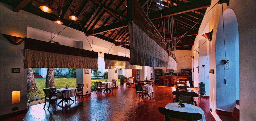 Colonial-style waterfront hotel in Kerala with traditional shuttered windows, wrap-around veranda, and palm trees against cloudy skies