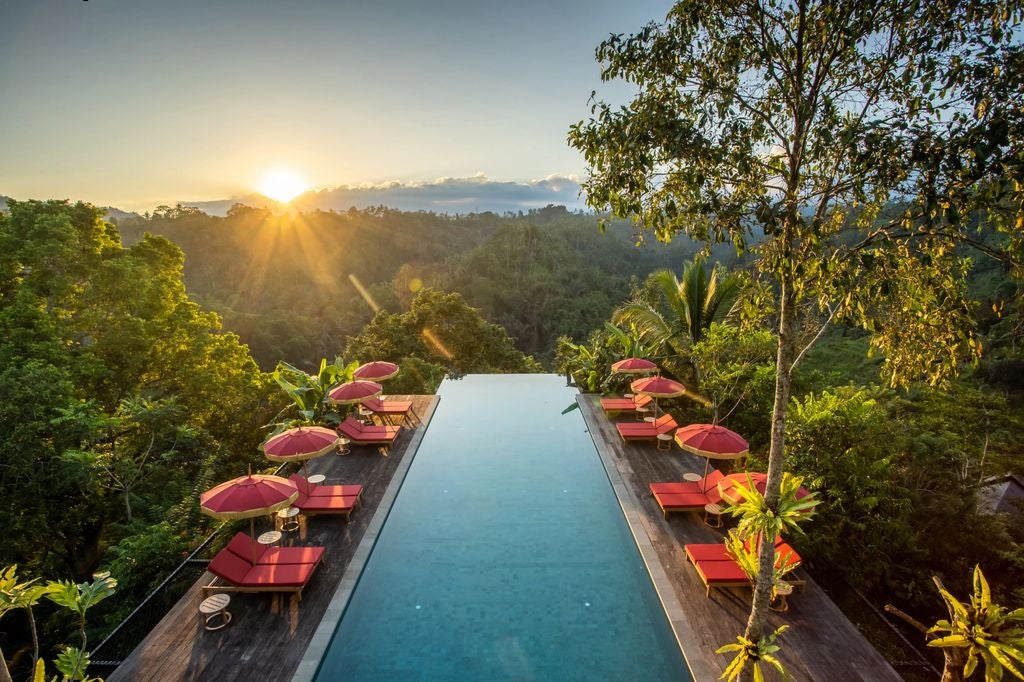 Open-air luxury villa nestled in Bali's jungle, featuring infinity pool overlooking lush green valley and distant mountains at sunset