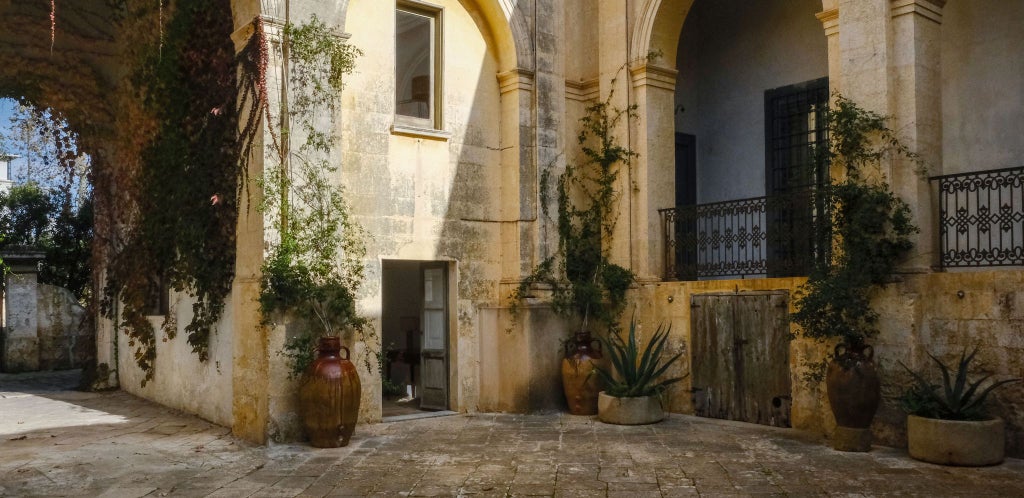 Elegant Italian luxury suite with high ceilings, antique white walls, ornate chandelier, and minimalist four-poster bed in historic Palazzo Daniele