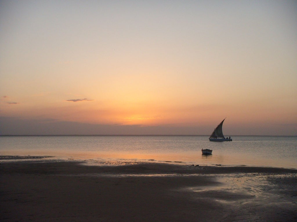 Palm-fringed white sand beach meets turquoise waters in Mozambique's Bazaruto Archipelago, with traditional dhow boat sailing offshore