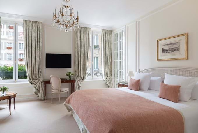 Elegant hotel room with queen bed, antique-style furniture, silk curtains and French doors opening to manicured garden views in Paris