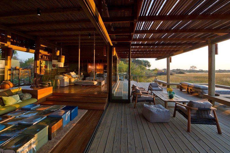 Elevated wooden walkway leads to luxurious tented suites overlooking Okavango Delta, with acacia trees and golden grasslands at sunset