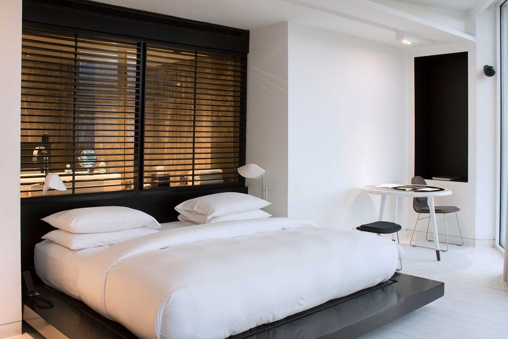 Modern minimalist corner hotel room with floor-to-ceiling windows, sleek gray furnishings, and panoramic city views of Monterrey's urban landscape at sunset.