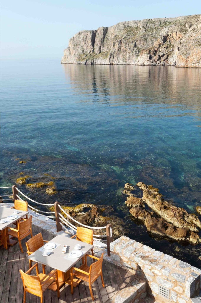 Stone and whitewashed traditional Greek seaside hotel with blue shutters nestled against rocky coastline, overlooking turquoise Aegean waters at scenic Scenset destination