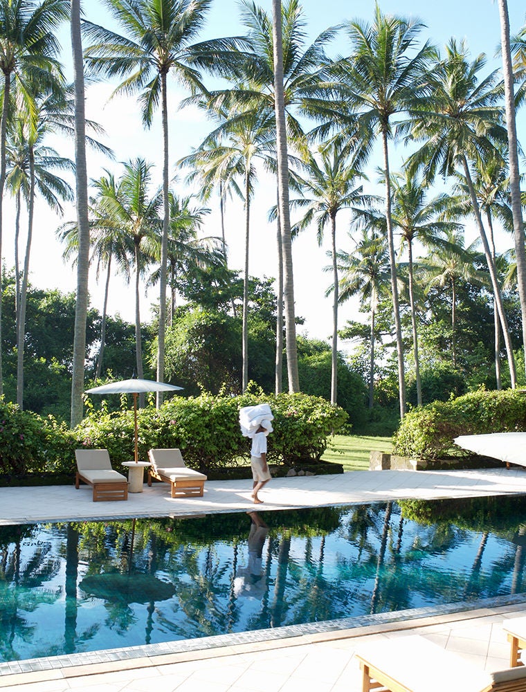 Three-tiered infinity pools cascade down a lush hillside overlooking the Bali Sea, with elegant palm trees and traditional thatched pavilions