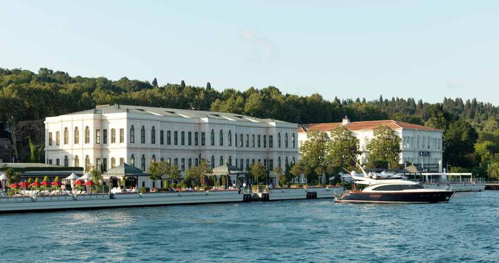 Historic luxury hotel with neoclassical white facade along Bosphorus strait, featuring waterfront terrace and manicured gardens at sunset