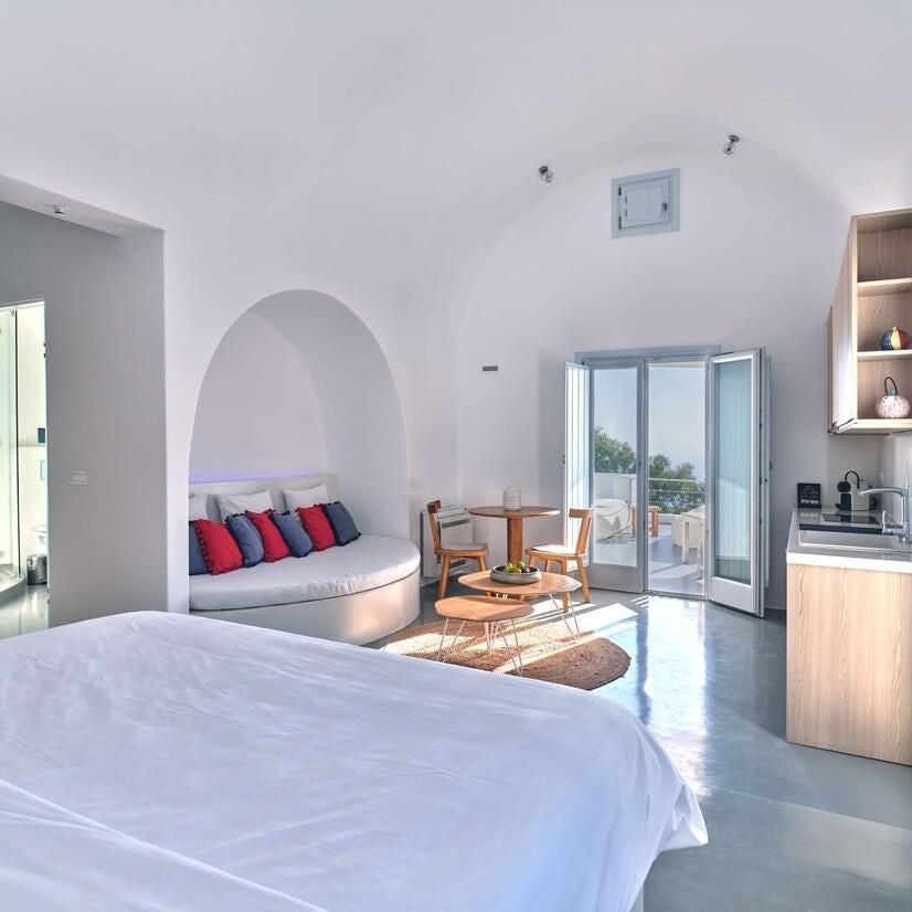 Luxury white-washed garden suite with panoramic Aegean Sea view, private terrace, blue-domed architecture, and lush Mediterranean greenery in Santorini, Greece