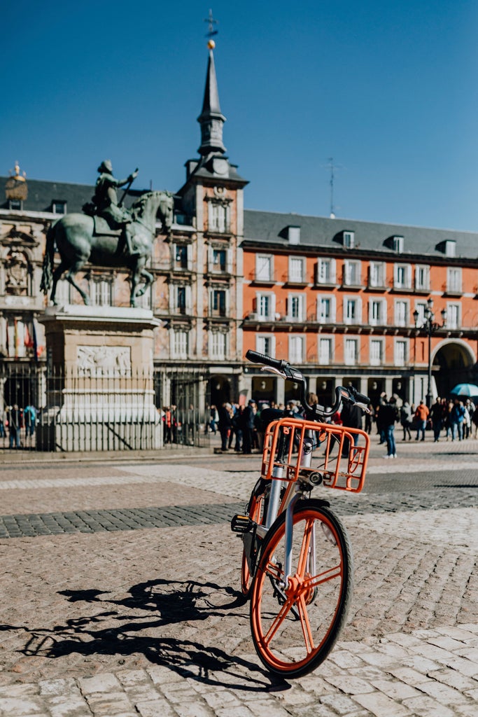 Cyclists riding through historic Madrid streets, passing elegant Baroque architecture, Royal Palace, and vibrant Plaza Mayor during golden afternoon light