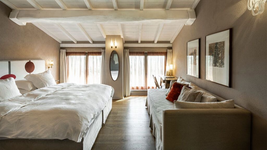 Elegant Tuscan-style hotel room with soft neutral tones, minimalist wooden furniture, and large windows overlooking lush olive grove landscape