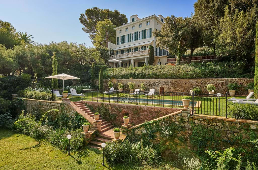 Iconic French Riviera luxury hotel with white facade and palm trees overlooking Mediterranean Sea from dramatic coastal cliffs