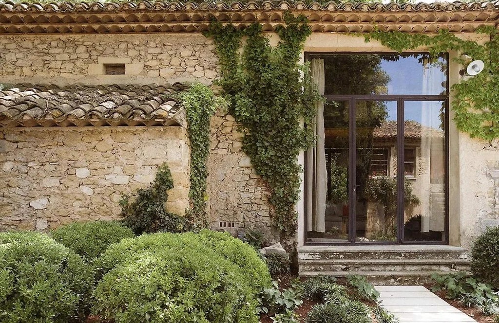 Elegant stone boutique hotel facade in Provence, with rustic warm-toned walls, lush green shutters, and soft Mediterranean sunlight casting gentle shadows