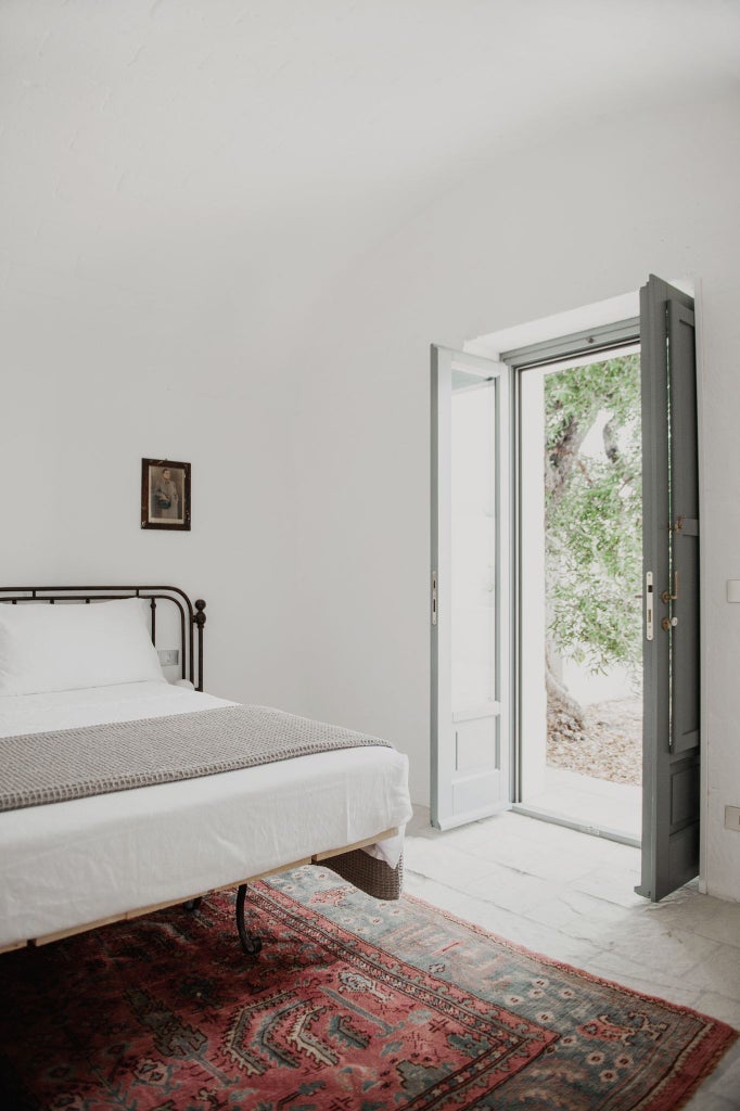 Minimalist white stone bedroom with rustic wooden furniture, soft natural light, overlooking an expansive olive grove landscape in scenic southern Italy