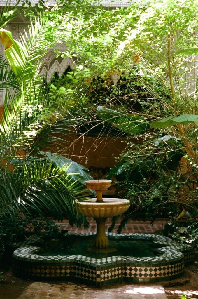 Luxurious Moroccan riad courtyard with pink walls, ornate archways, lush palm trees and traditional tiled fountain in shaded garden