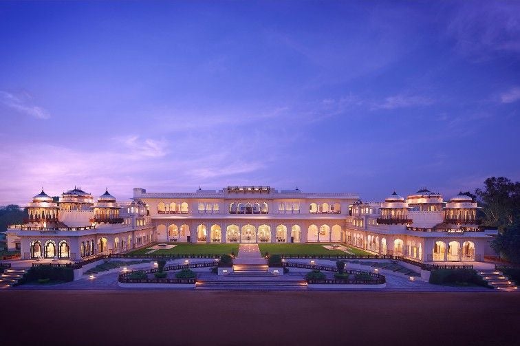 Ornate sandstone facade of Rambagh Palace hotel with grand archways, manicured gardens, and traditional Rajasthani architecture at sunset