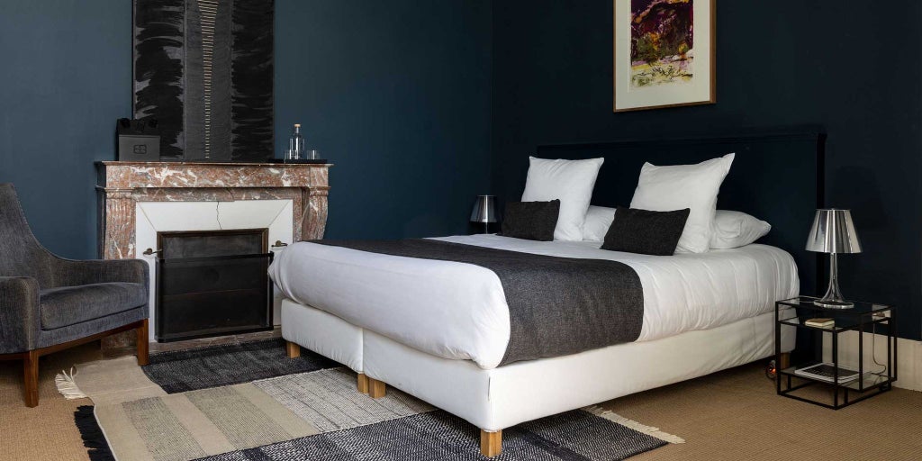 Elegant minimalist hotel room with exposed stone walls, crisp white linens, contemporary design, and soft natural light streaming through large windows overlooking French countryside