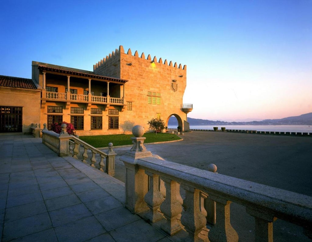 Historic stone castle-hotel Parador de Baiona perched on rocky peninsula, overlooking blue Atlantic Ocean with luxury boats in harbor