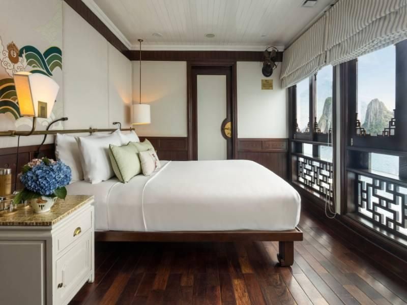 Elegant Vietnamese luxury cabin interior with wooden furnishings, plush white bedding, panoramic window overlooking emerald waters of Halong Bay