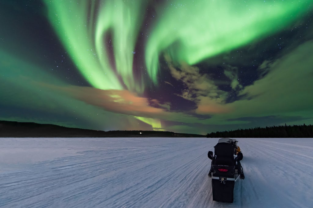 Luxury snowmobile expedition racing across pristine Finnish snowscape under vibrant green and blue Northern Lights dancing across starry Arctic night sky