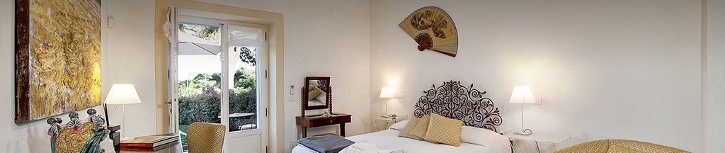 Elegant Sicilian hotel room with soft ivory linens, minimalist wooden furniture, and arched window overlooking lush Mediterranean garden at Donna Coraly Resort.