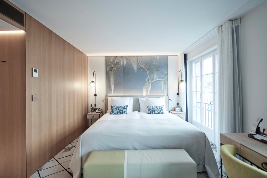 Elegant riverside hotel room with plush white bedding, blue accent walls, crystal chandelier, and panoramic windows overlooking Zurich's waterfront