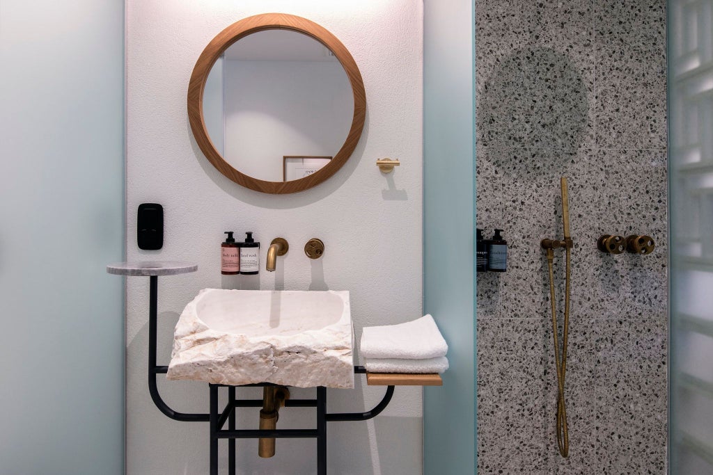 Elegant minimalist hotel room with stone walls, wooden floor, plush king bed, large window overlooking Athenian cityscape, modern luxury design.