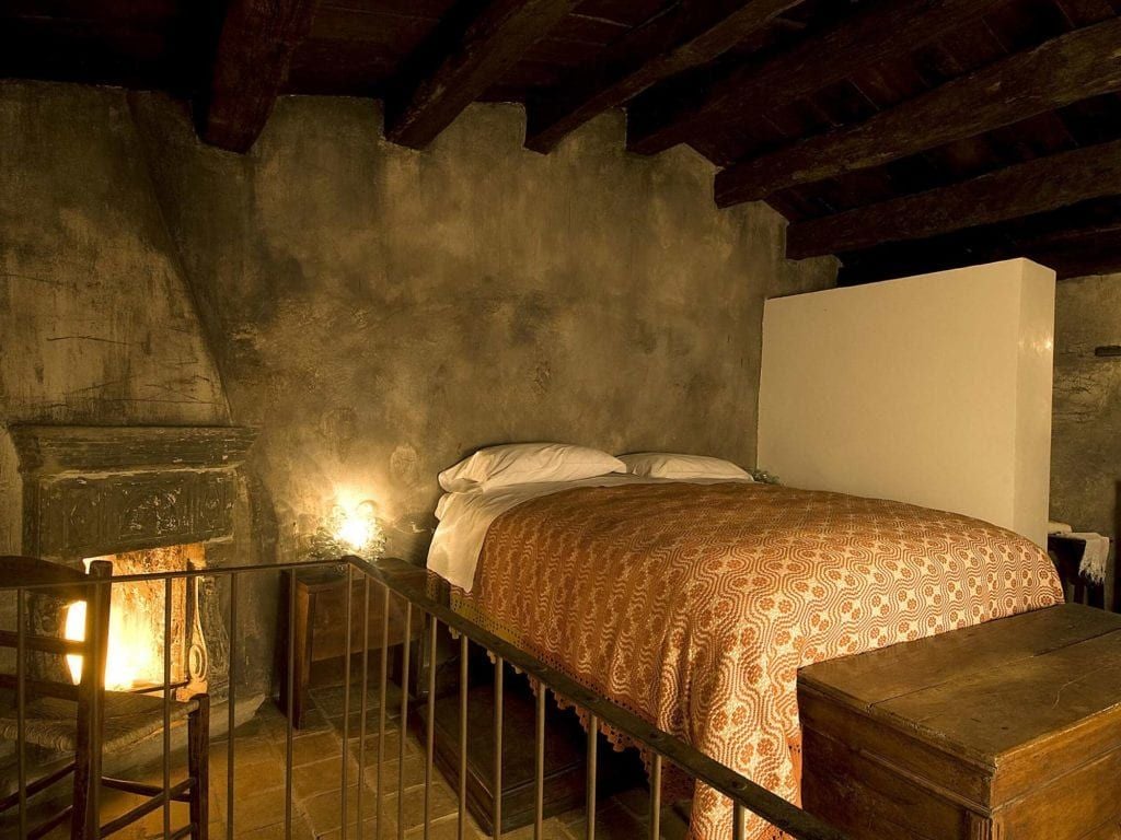 Rustic Italian stone hotel room with antique wooden bed, soft cream linens, exposed wooden beams, and panoramic window overlooking historic village rooftops