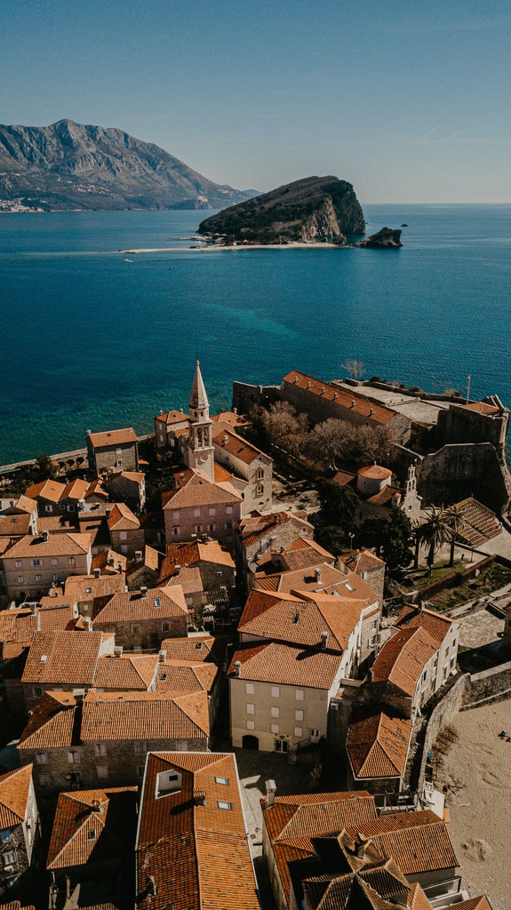 Traditional stone village buildings nestled on green mountainside with dramatic limestone cliffs rising above Bay of Kotor at sunset