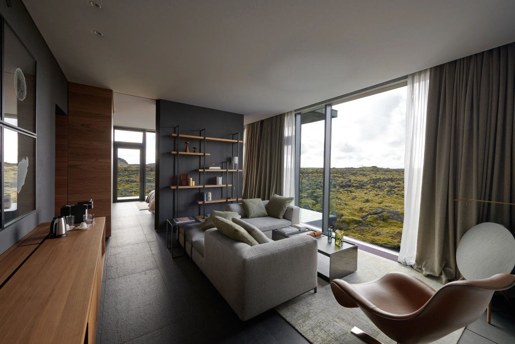 Luxurious minimalist Lagoon Suite with floor-to-ceiling windows overlooking Iceland's volcanic landscape, featuring elegant gray decor and natural stone accents.