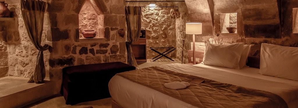 Elegant stone-walled junior suite with plush white bedding, rustic wooden furnishings, and expansive view of Cappadocia's unique volcanic landscape