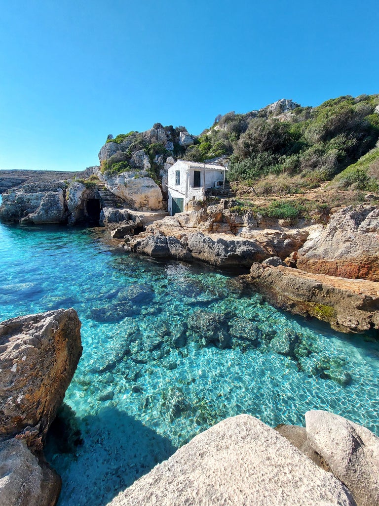 Mediterranean turquoise waters meet rugged limestone cliffs along Menorca's pristine coastline, with whitewashed villas nestled in the hills