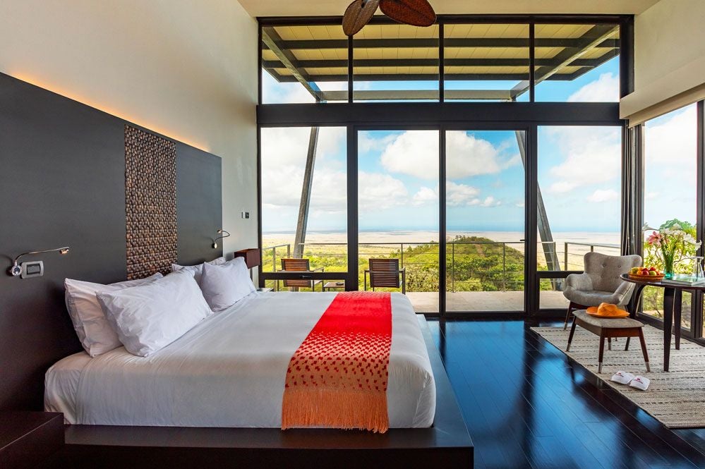 Spacious balcony overlooking lush Galapagos landscape, with modern minimalist room interior, crisp white linens, and floor-to-ceiling glass doors at Pikaia Lodge