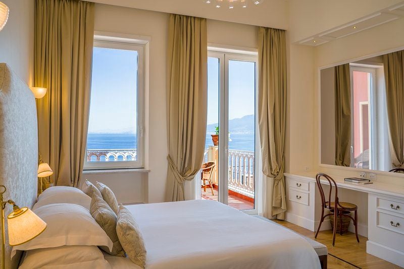 Elegant hotel balcony with ornate railing overlooks Bay of Naples, featuring antique furnishings and Mediterranean architecture details