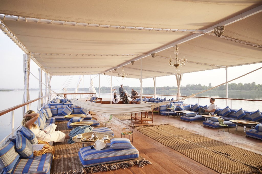 Traditional wooden dahabiya boat with white canopied deck gliding along the Nile, featuring elegant cream sails and ornate Arabic design details