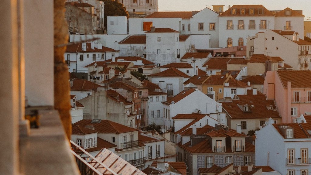 Aerial cityscape of Lisbon's historic pastel buildings, terracotta roofs, and winding streets leading to the waterfront against sunset sky