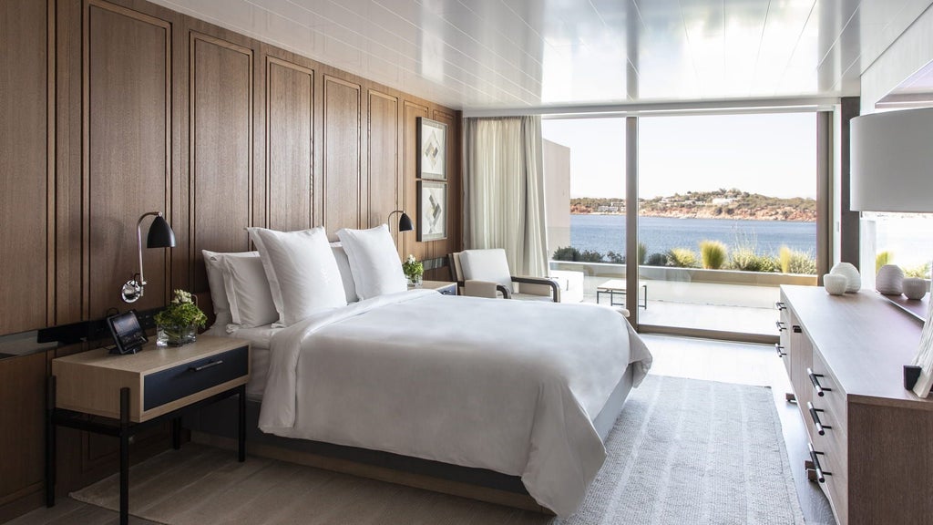 Luxurious hotel suite with floor-to-ceiling windows overlooking Aegean Sea, featuring modern furniture and private terrace with lounge chairs