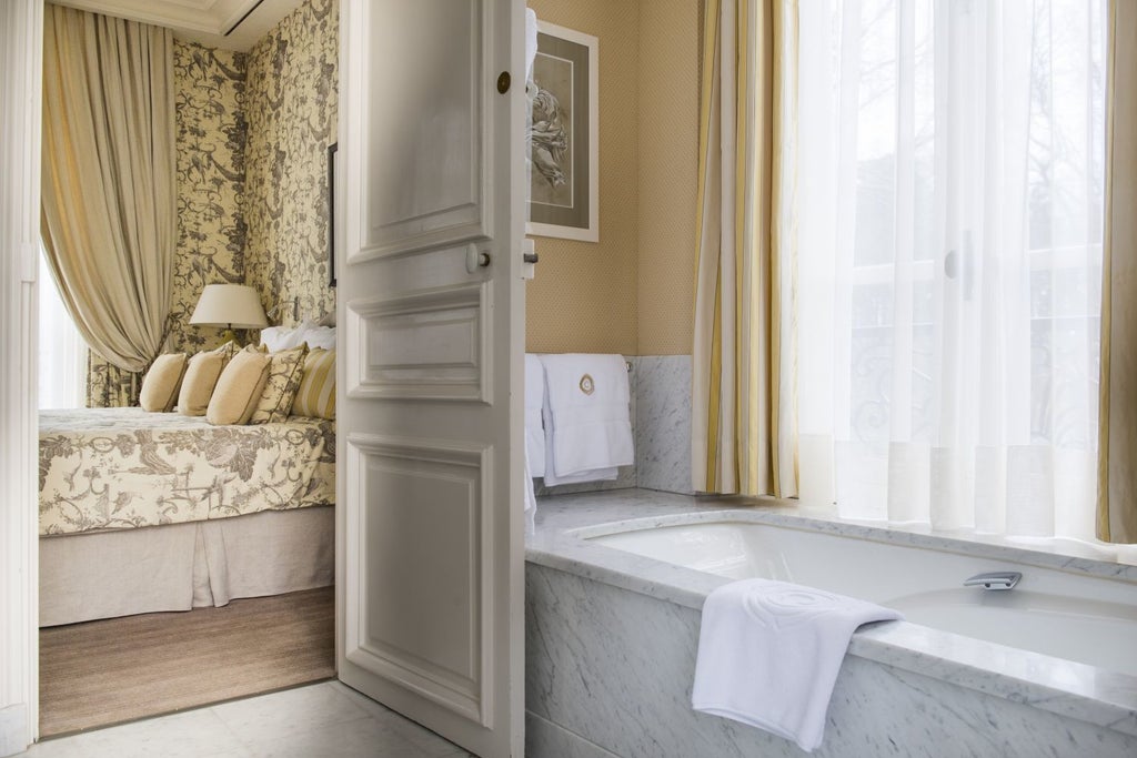 Ornate French hotel room with crystal chandelier, antique furnishings, gold-framed artwork, and floor-to-ceiling windows with garden views