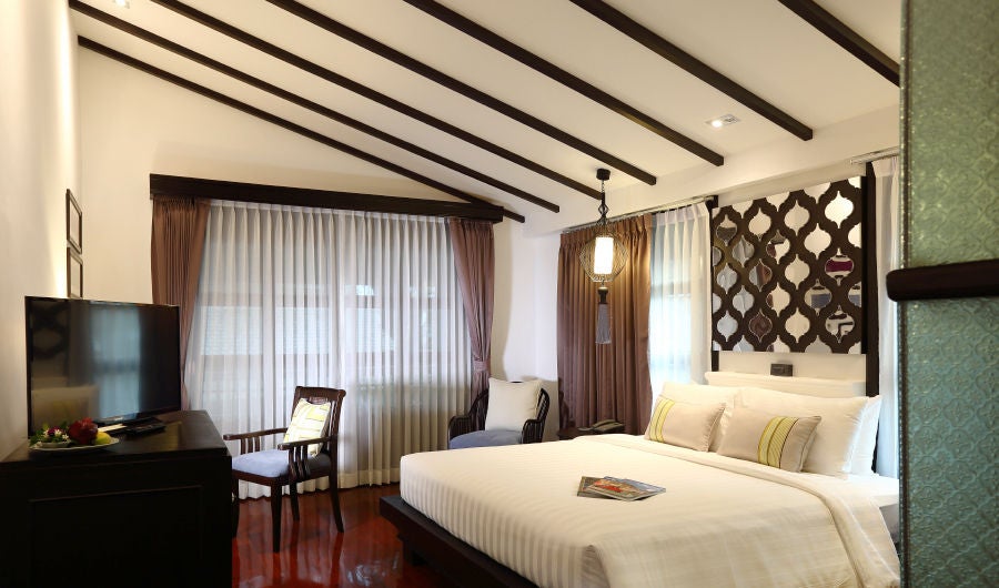 Traditional Thai-style luxury suite with elegant wooden furnishings, king bed, white linens, and ornate local textiles against dark wood floors