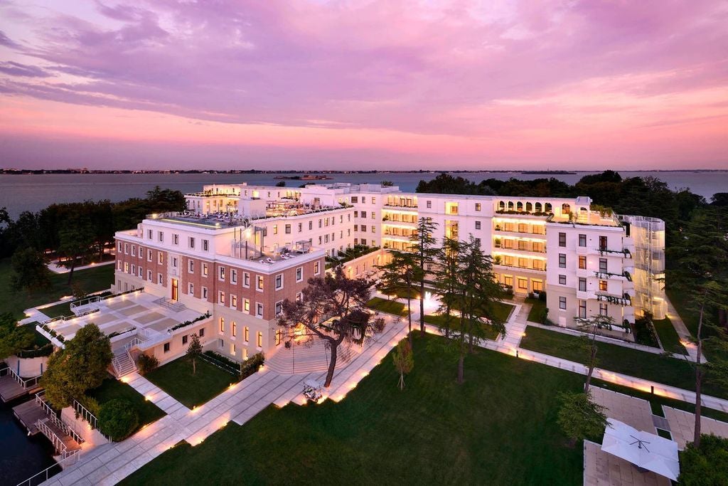 White marble-clad luxury hotel on private Venetian island, featuring elegant archways and manicured gardens overlooking lagoon at sunset