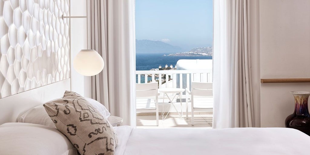 Luxurious white-washed Cycladic hotel room with blue accents, private balcony overlooking Mykonos' azure waters and pristine landscape at sunset